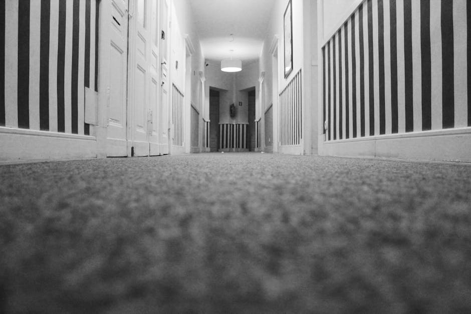 A wide-angle view of a corridor with a low perspective, showing a textured carpet with a mottled pattern on the floor, flanked by white wooden door panels and decorative striped wallpaper in shades of