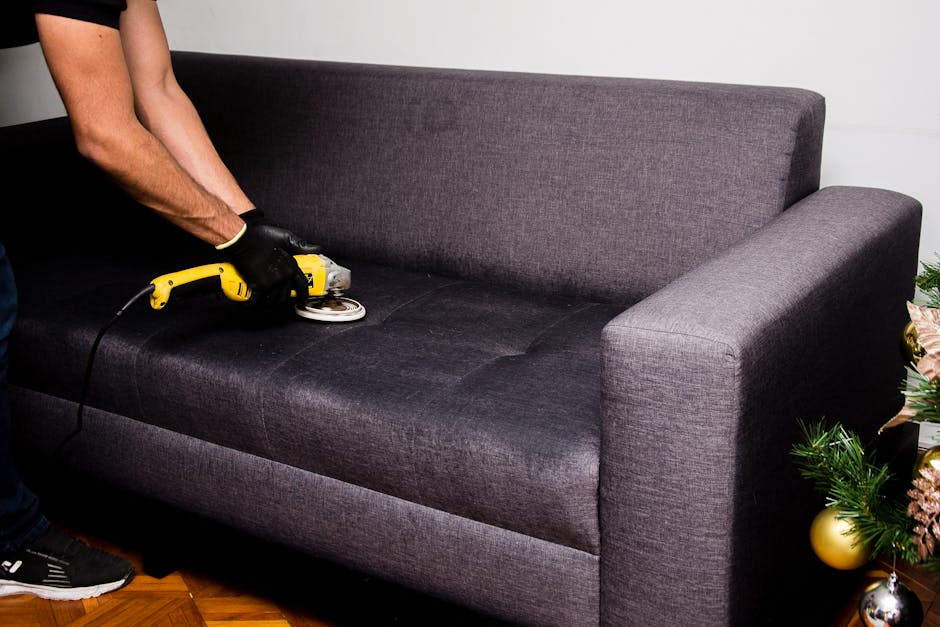 A person wearing black gloves is using a yellow handheld upholstery cleaning machine on a dark grey fabric sofa in a well-lit living room. The sofa has a high backrest and wide armrests, and is situat