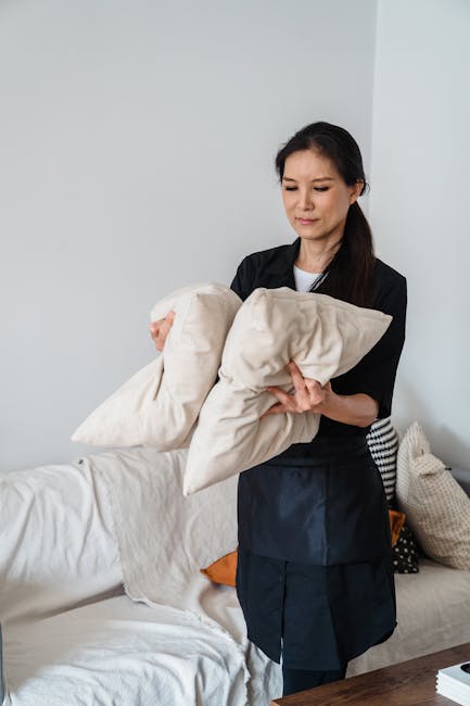 A person wearing black gloves is using a yellow handheld upholstery cleaning machine on a dark grey fabric sofa in a well-lit living room. The sofa has a high backrest and wide armrests, and is situat